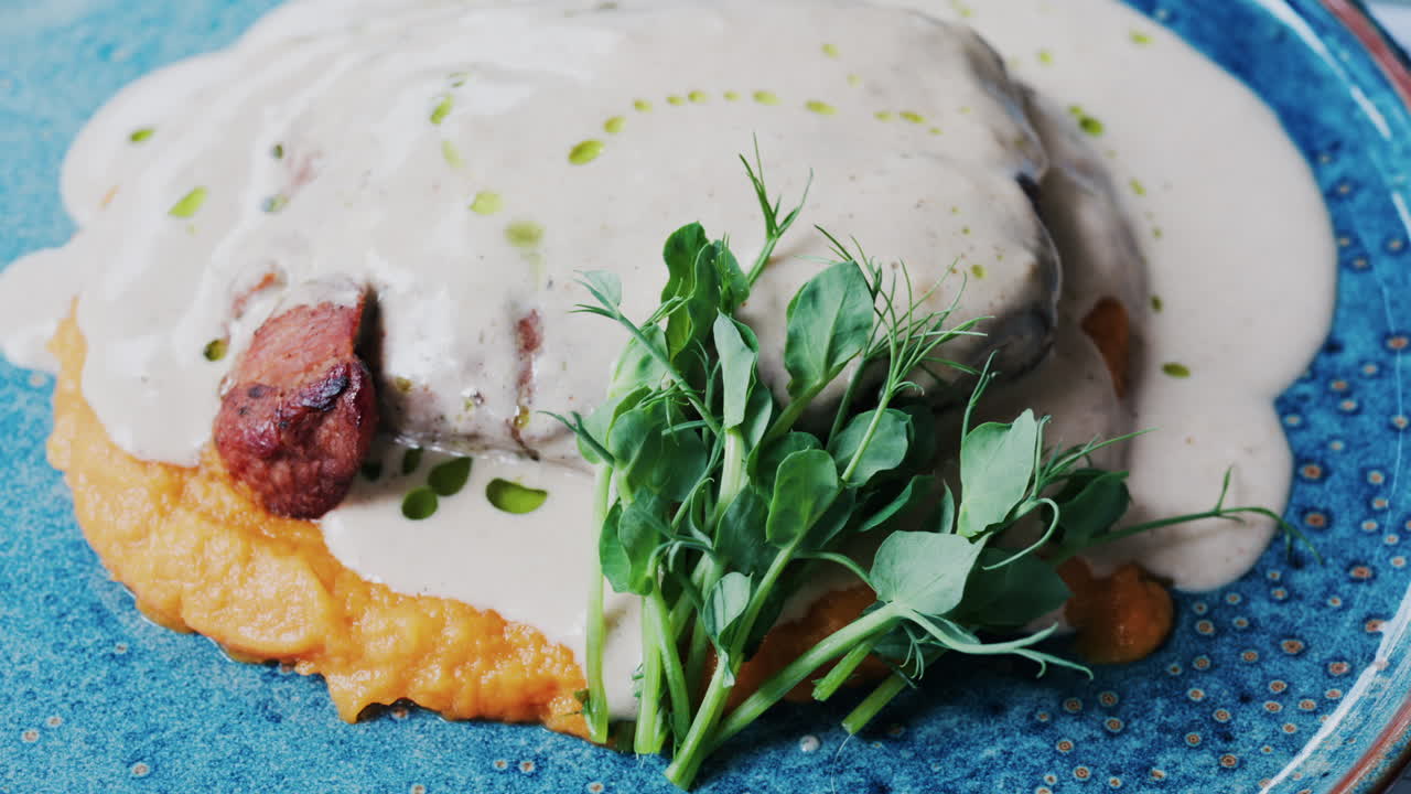Close-up of a piece of grilled meat covered in a creamy white sauce, accompanied by a vibrant orange puree base and garnished with fresh green herbs