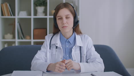 una médica jubilada está consultando en línea mirando a la cámara y asintiendo con la cabeza escuchando por auriculares servicio de telemedicina