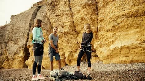 A-blonde-girl-in-a-blue-jacket,-a-blonde-girl-with-curly-hair-in-black-clothes-and-a-brunette-guy-in-a-gray-T-shirt-put-on-a-belay-before-they-start-rock-climbing-on-a-rocky-shore-near-the-yellow-rocks-during-the-day