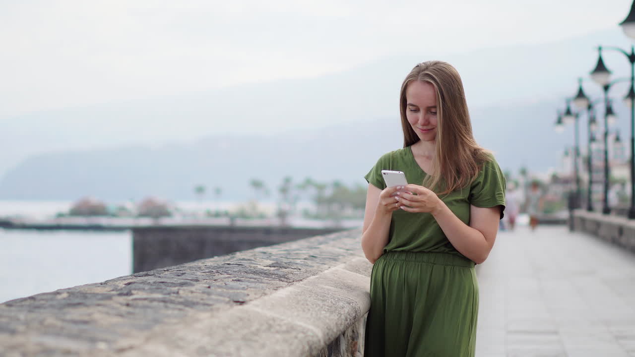 Against the beach's seascape horizon, a traveler employs a mobile phone in her hand. Her gaze rests upon the azure ocean and radiant sun, encapsulating a dynamic summer lifestyle
