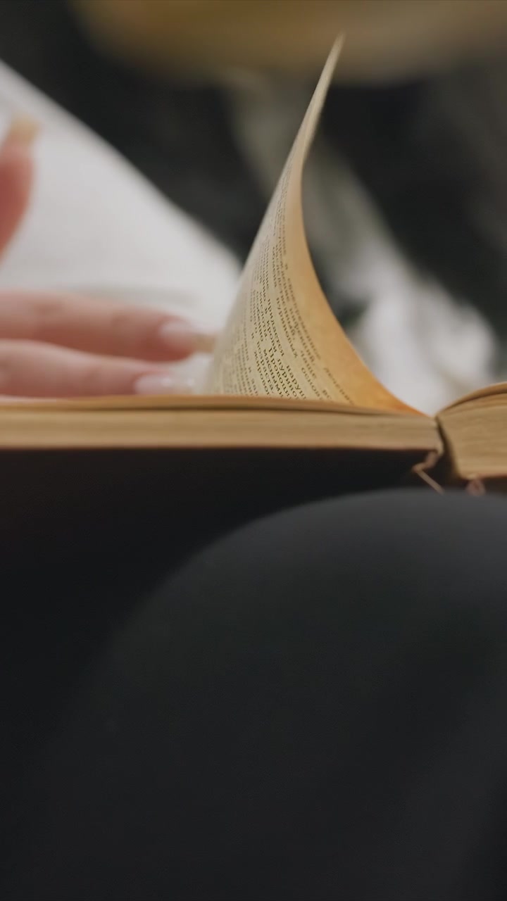 Mani che sfogliano testi su carte invecchiate. Forme e colori indistinti riempiono l'area sfocata. Una vista ravvicinata dei libri sfogliati rivela un ambiente interno dettagliato