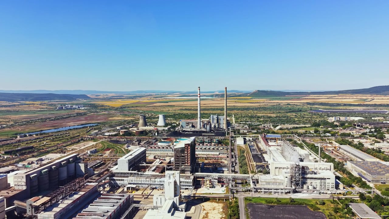 Area industriale che mostra grandi fabbriche e torri di raffreddamento con tempo sereno, campi verdi e colline sullo sfondo vicino a un fiume.