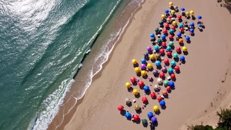 aerial view of a sandy beach with a mosaic of colorful beach umbrellas next to the ocean waves creating a vibrant and cheerful summer scene