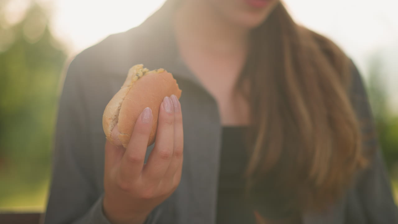 빛이 비치는 야외 환경에서 책을 읽는 동안 샤우마를 들고 회색 을 입은 여자의 클로즈업, 부드러운 빛은 그녀의 손과 간식을 밝히고, 초록색과 따뜻한 빛의 흐릿한 배경으로