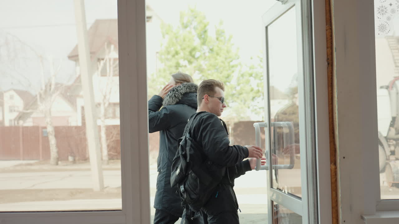 Man with tattoo on neck adjusting sunglasses while another man in winter coat walking out perspective of two individuals at entrance, outdoor houses and street visible through glass windows