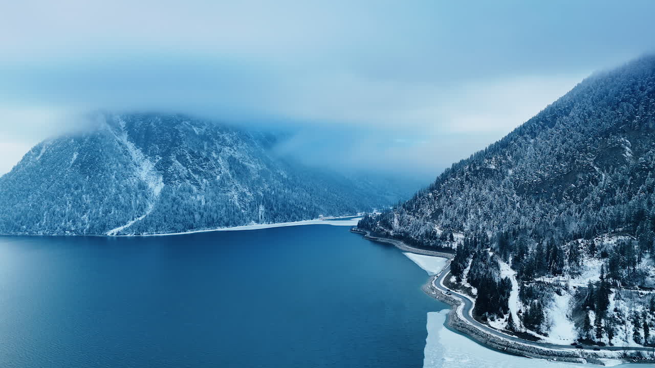 A highway built on the lakefront of the waterscape surrounded by splendid rocks. Stunning aerial scenery of mountains covered with pine tree forests and thick cloudscape at the tops.