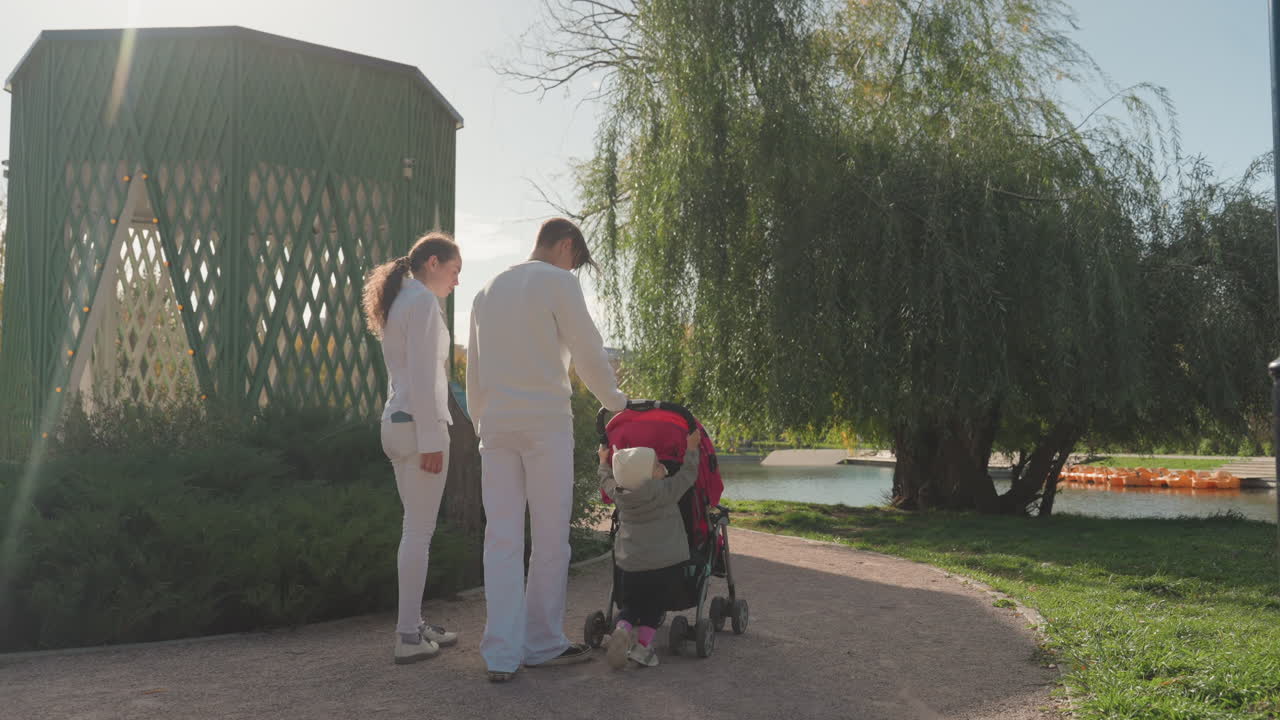 Coppia bianca con passeggino in riva al lago, giovani genitori sposati si fermano vicino a un gazebo e un salice sotto una calda luce solare, controllando il bambino nel passeggino, tenera conversazione, sereno momento familiare durante una passeggiata pomeridiana