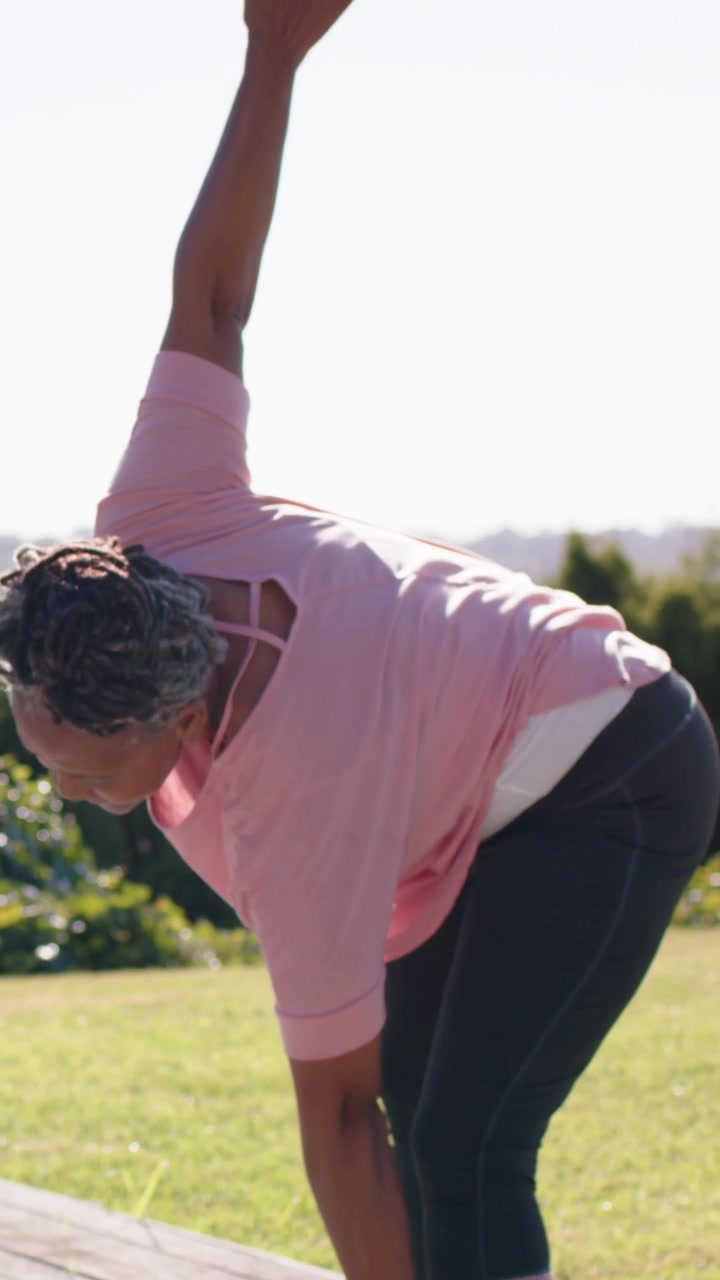 Video verticale: Donna con top rosa e orologio, che alza il braccio destro e si piega per la salute su un tappetino da yoga, spazio per il testo.