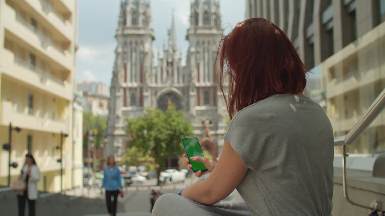 도시 거리에 앉아있는 녹색 화면의 휴대전화를 사용하는 젊은 여성, 어깨 뷰. 거리에서 chromakey와 함께 휴대전화를 스크롤하는 여성.