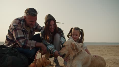 Close-up-shot-of-a-happy-cream-colored-dog-resting-on-a-deserted-seashore-with-its-owners.-A-happy-brunette-man-and-his-wife-in-a-green-checkered-shirt-are-stroking-their-dog-and-their-little-daughter-is-sitting-near-them-during-a-picnic-and-hike-in-the-summer