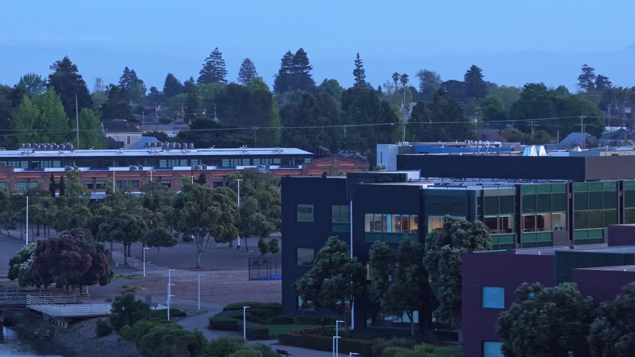 Scenic aerial view flying over the waterfront of Encinal Basin in Alameda, California, revealing modern buildings, a public park, and lush trees during a quiet blue hour evening