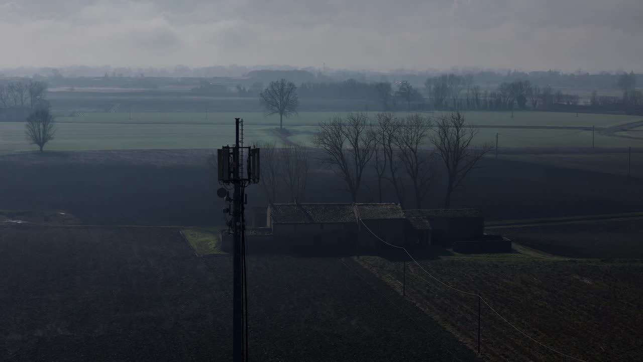 Drone pushes in toward communication tower in foggy farmland near Folgarole—revealing antennas, leafless trees, and rural textures in Piacenza’s quiet Emilia-Romagna countryside