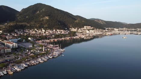 Vista-Aérea-Cinematográfica-De-La-Ciudad-De-Fethiye-Con-Barcos-Estacionados-En-La-Costa-Mediterránea-Y-Montañas-Al-Fondo,-Turquía.