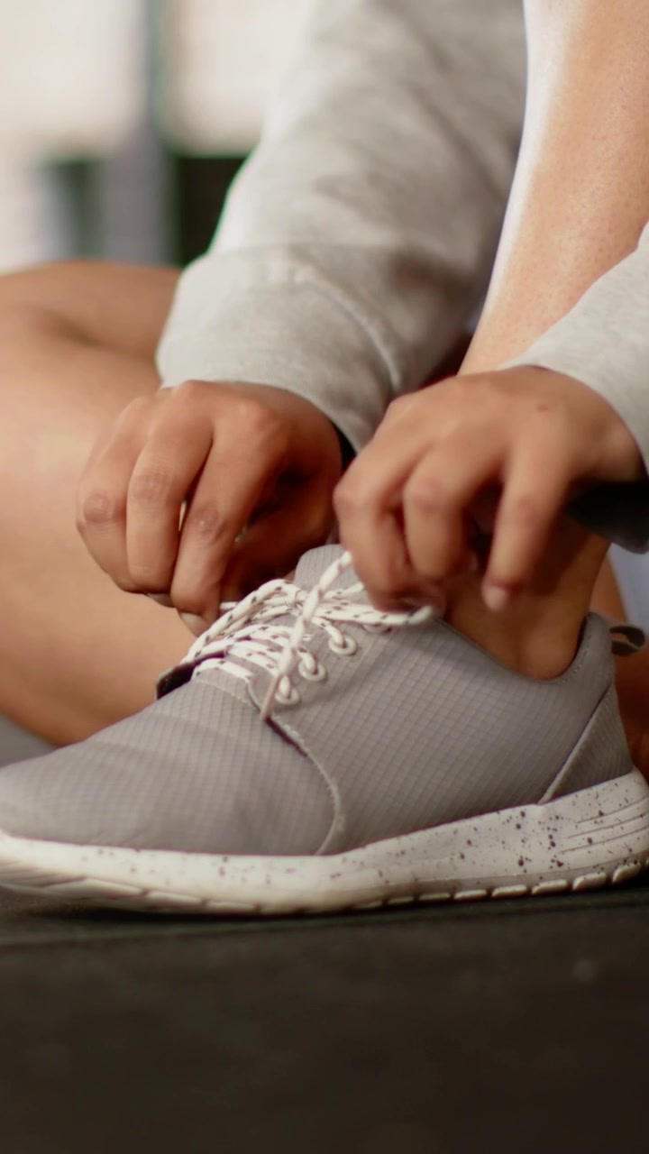Video verticale: Donna in pantaloncini che si prepara per l'allenamento, allacciando i lacci fantasia su una sneaker grigia in palestra.