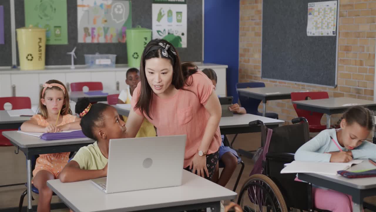 Felice insegnante di diverse etnie che aiuta una bambina a usare un laptop in una classe di scuola elementare, in slow motion.