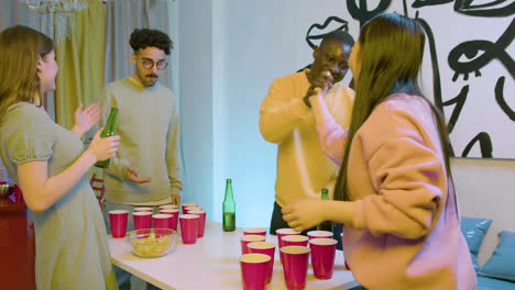 happy multiethnic young friends playing beer pong at home