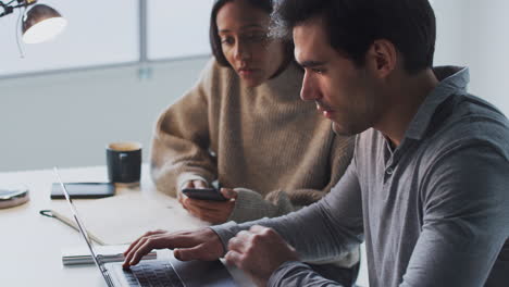 Businessman-Working-On-Laptop-At-Desk-Collaborating-With-Female-Colleague-Using-Mobile-Phone