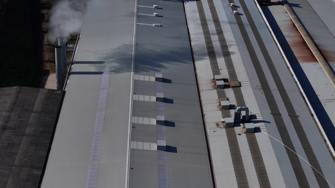 Industrial facility rooftop releases a white steam plume from an exhaust stack above production buildings with ventilation equipment and skylights, drone orbits