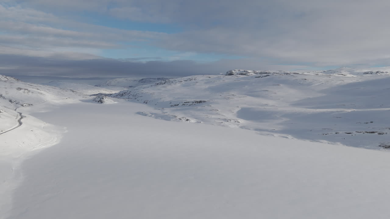 Sideways flying wide landscape droneshot flying near a frozen lake in Norway on a bright day with the sun in the background and a single road underneath LOG