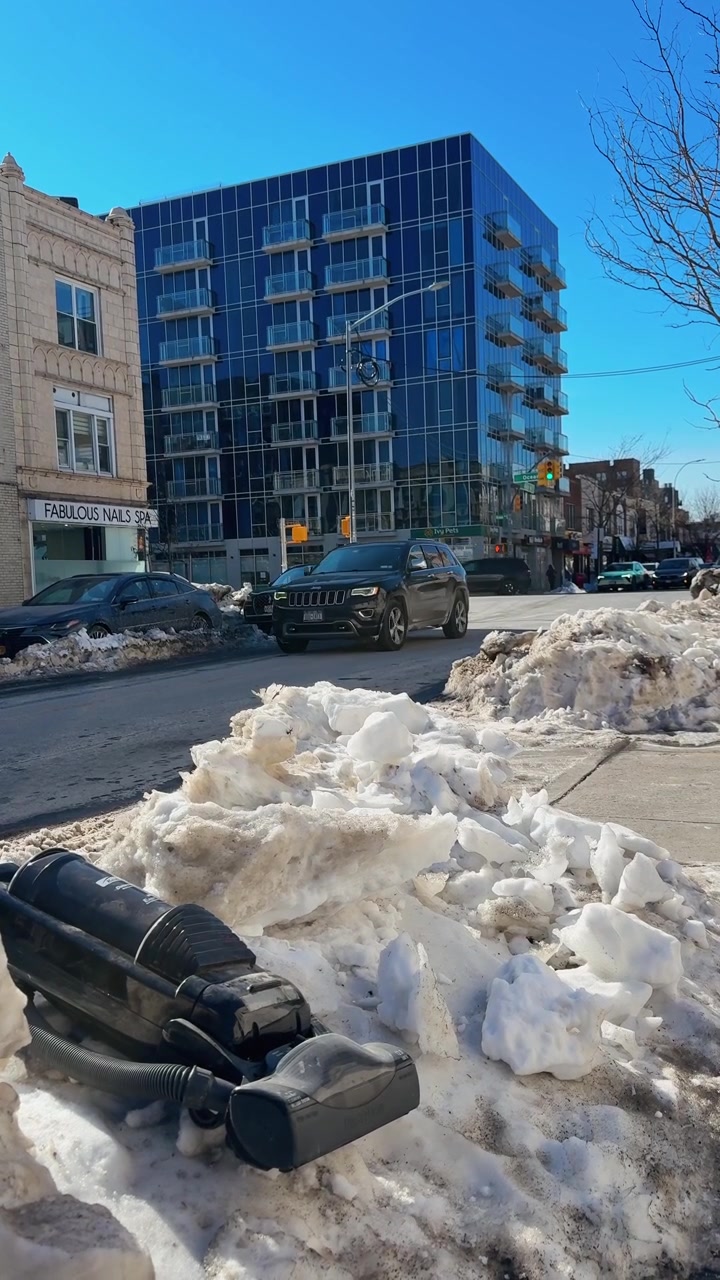 Nueva York, EE. UU., 25 de enero de 2026: Aspiradora negra abandonada en un montón de nieve en una calle de la ciudad. Aspiradora negra rota tirada en un banco de nieve sucio cerca de un moderno edificio de cristal y un salón de uñas.