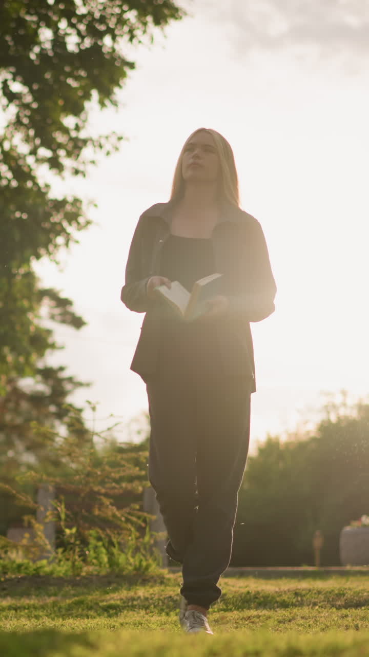 회색 옷을 입은 여자가 책 읽는 동안 골든 아워 동안 야외에서 풀을 고, 빛이 주차된 차와 나무의 전망과 함께 주변을 조명합니다.