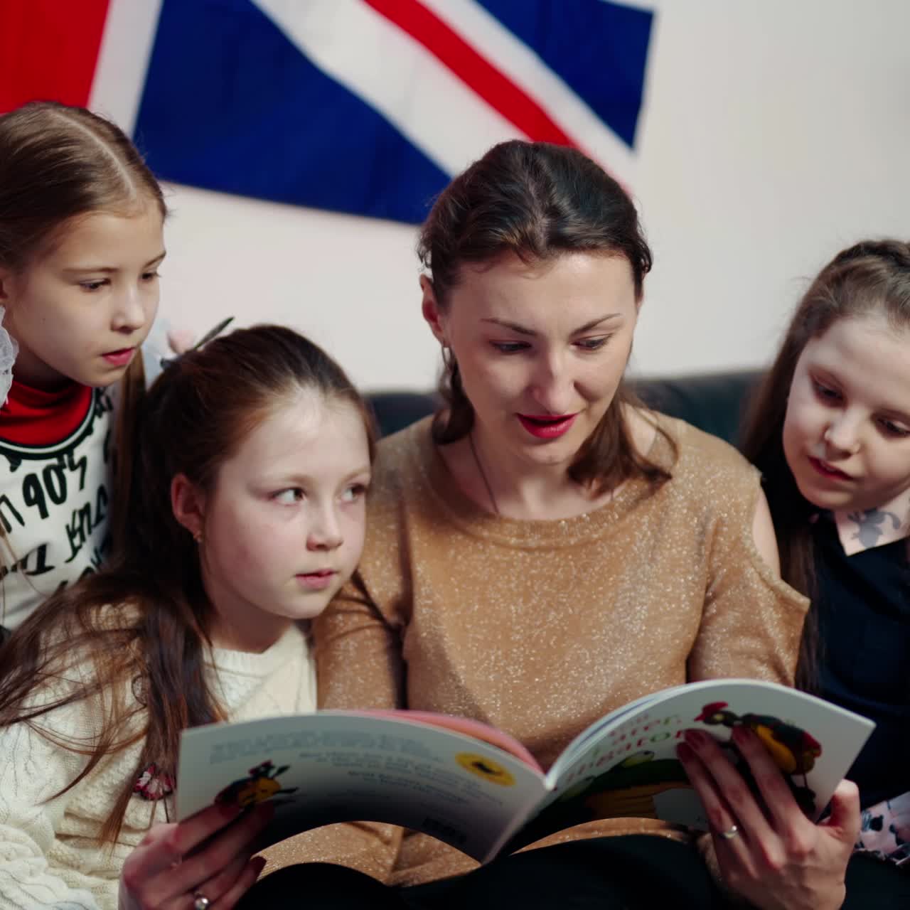 Teacher and schoolgirls on the sofa in class. Young female teacher reading a book to children while sitting together on the couch at school. Elementary education concept.