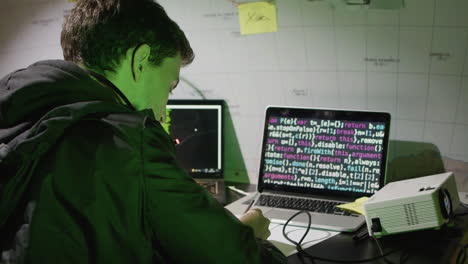 back view of a hacker at late night typing code on computer, making notes on paper while stealing network database data