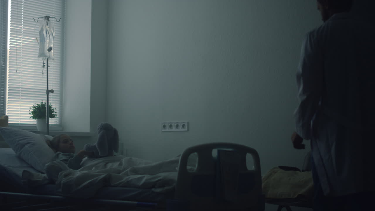 병원 침대에 누워있는 어린 소녀가 플러시 장난감으로 놀고 있습니다. 의사는 병실에서 환자를 방문합니다.