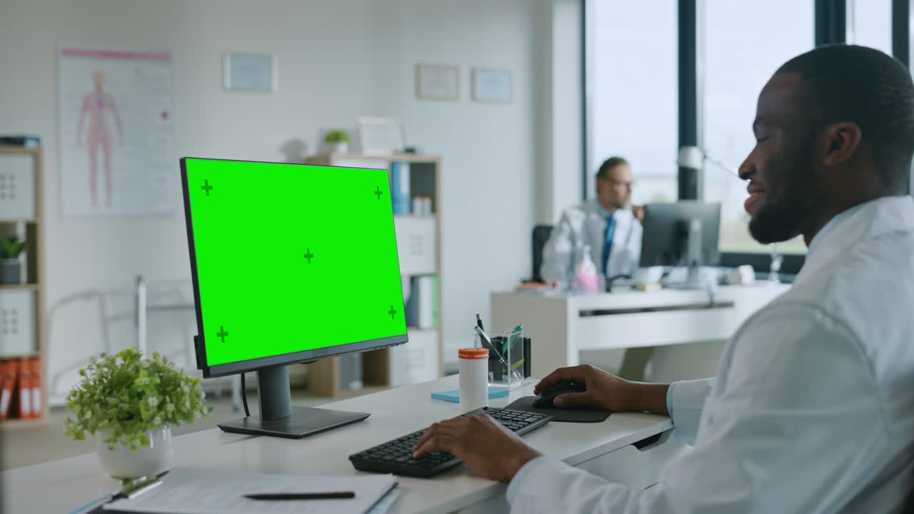 un medico afroamericano sta lavorando su un computer con schermo verde in una clinica sanitaria. un medico in camicetta bianca sta leggendo la storia medica dietro una scrivania nell'ufficio dell'ospedale.