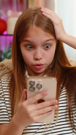child girl kid reads shocking message on smartphone with wide open eyes while sitting on home sofa