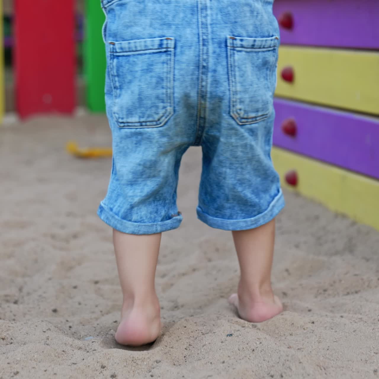 Lower part of a baby boy stepping by the sand on the playground. Little toddler feet come up to the colorful stairs