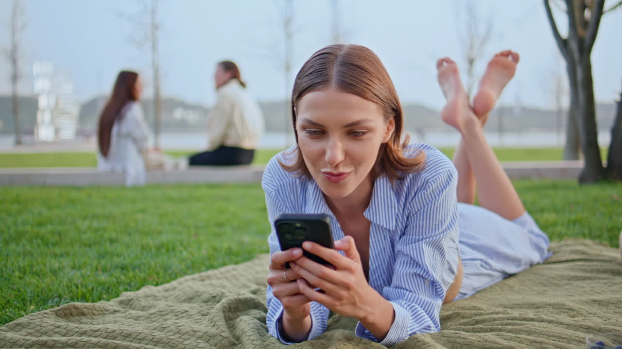 Girl messaging lying picnic blanket at green lawn closeup. Smiling woman texting