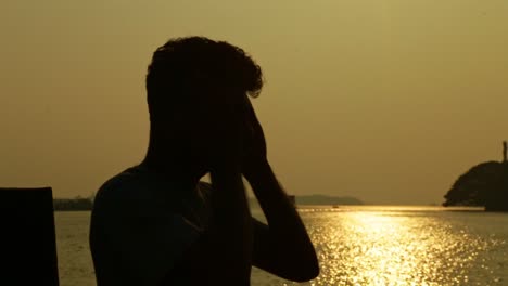 Hombre-Indio-Acariciando-Su-Rostro,-Hora-Del-Atardecer,-Casas-Flotantes-En-El-Fondo,-Silueta