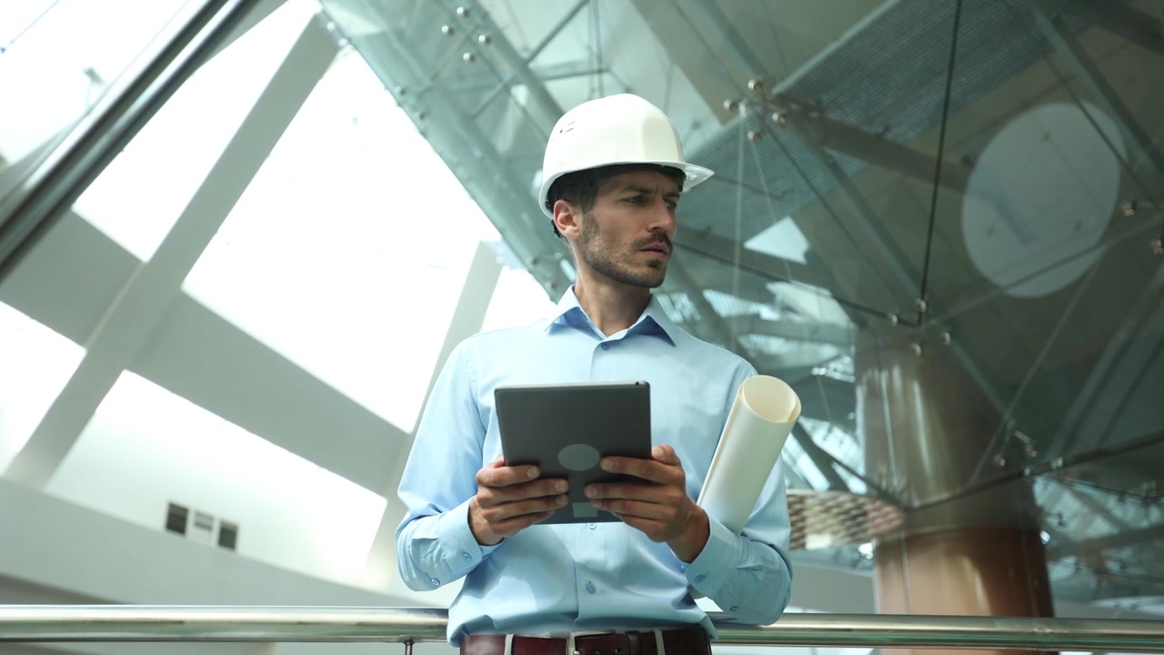 Ingeniero con sombrero usando una tablet y un plano en un edificio moderno