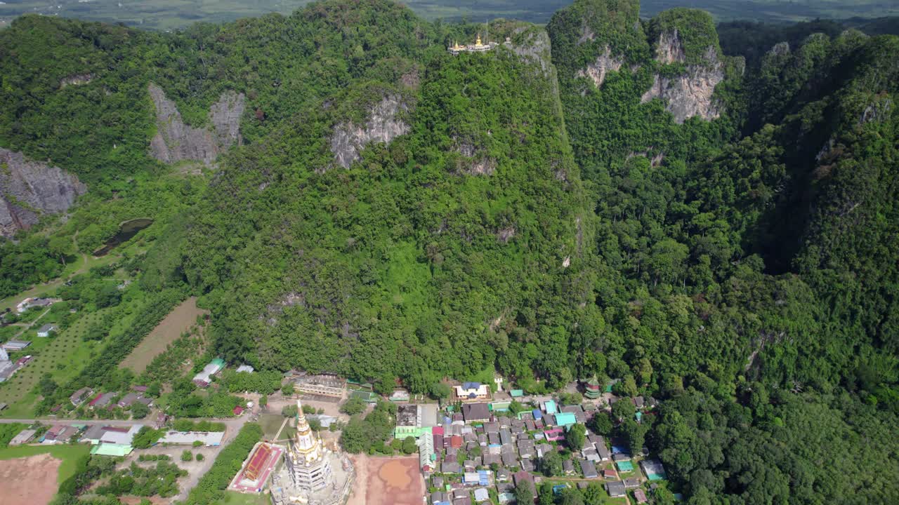 Cảnh quay drone nghiêng lên hé lộ Chùa Hang Hổ tọa lạc trên một ngọn núi đá vôi ở Krabi, Thái Lan, với rừng rậm và các công trình làng mạc hiển thị bên dưới, ghi lại địa điểm tôn giáo và phong cảnh