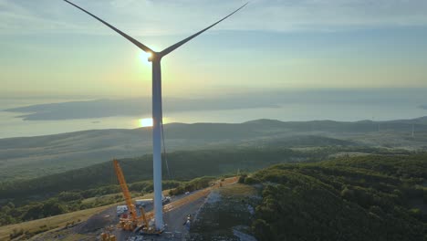 Installation-of-wind-turbine-crawler-crane-working-at-wind-tower-erection,-aerial-view-at-sunset-of-windmill-installed-on-top-of-hill-with-scenic-ocean-landscape