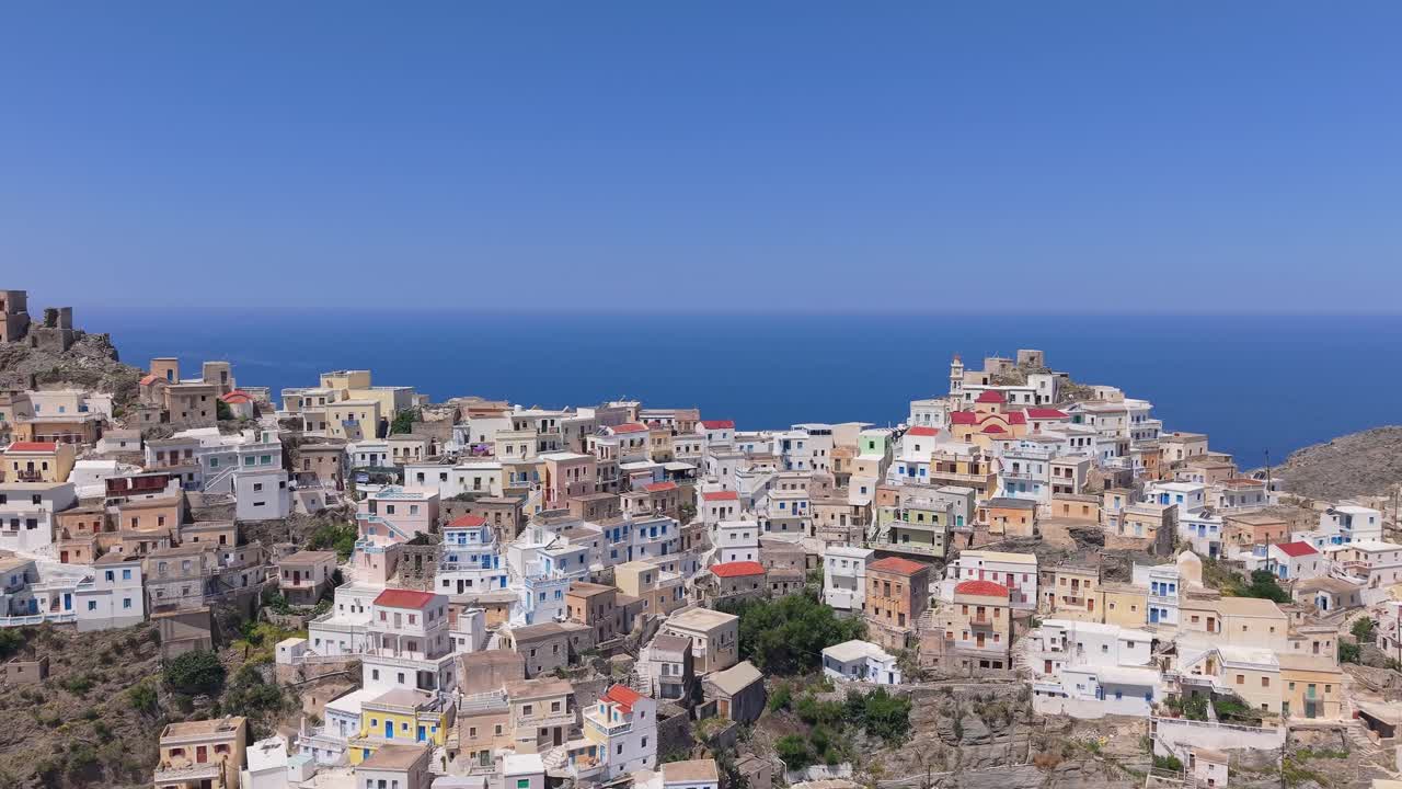 Drone shot approaching Olympos village in Karpathos, revealing the traditional houses and hillside as the Aegean Sea appears behind the village, highlighting stunning island scenery