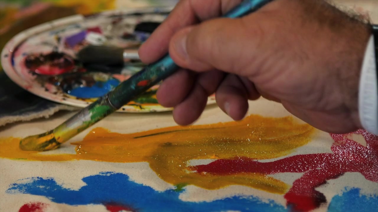 Primo piano di un artista che applica pittura acrilica gialla con un pennello su una tela astratta con pennellate colorate in uno studio d'arte creativo.