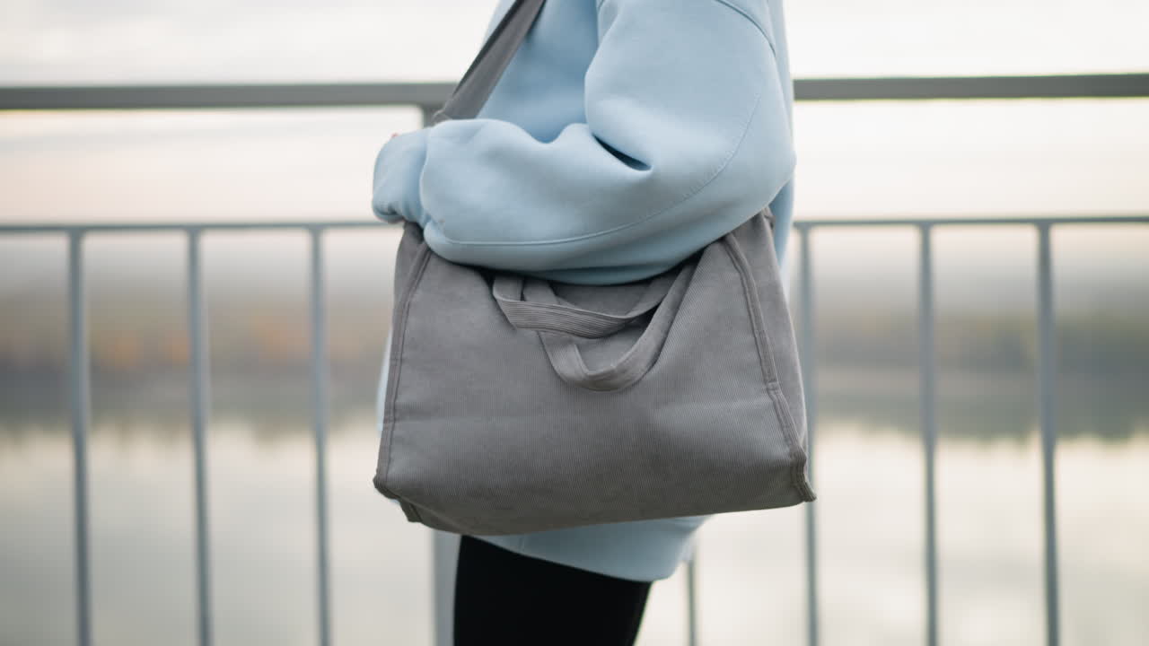 Close-up view of young woman holding a handbag while walking along an iron fence, background features tranquil river view, focus on her confident stroll in casual attire