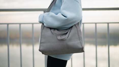close-up view of young woman holding a handbag while walking along an iron fence, background features tranquil river view, focus on her confident stroll in casual attire