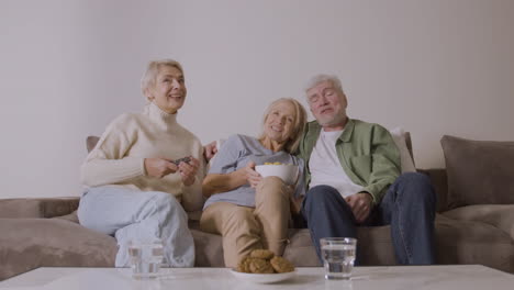 grupo de tres personas mayores viendo televisión, hablando y comiendo palomitas de caramelo mientras se sientan en el sofá en casa 3