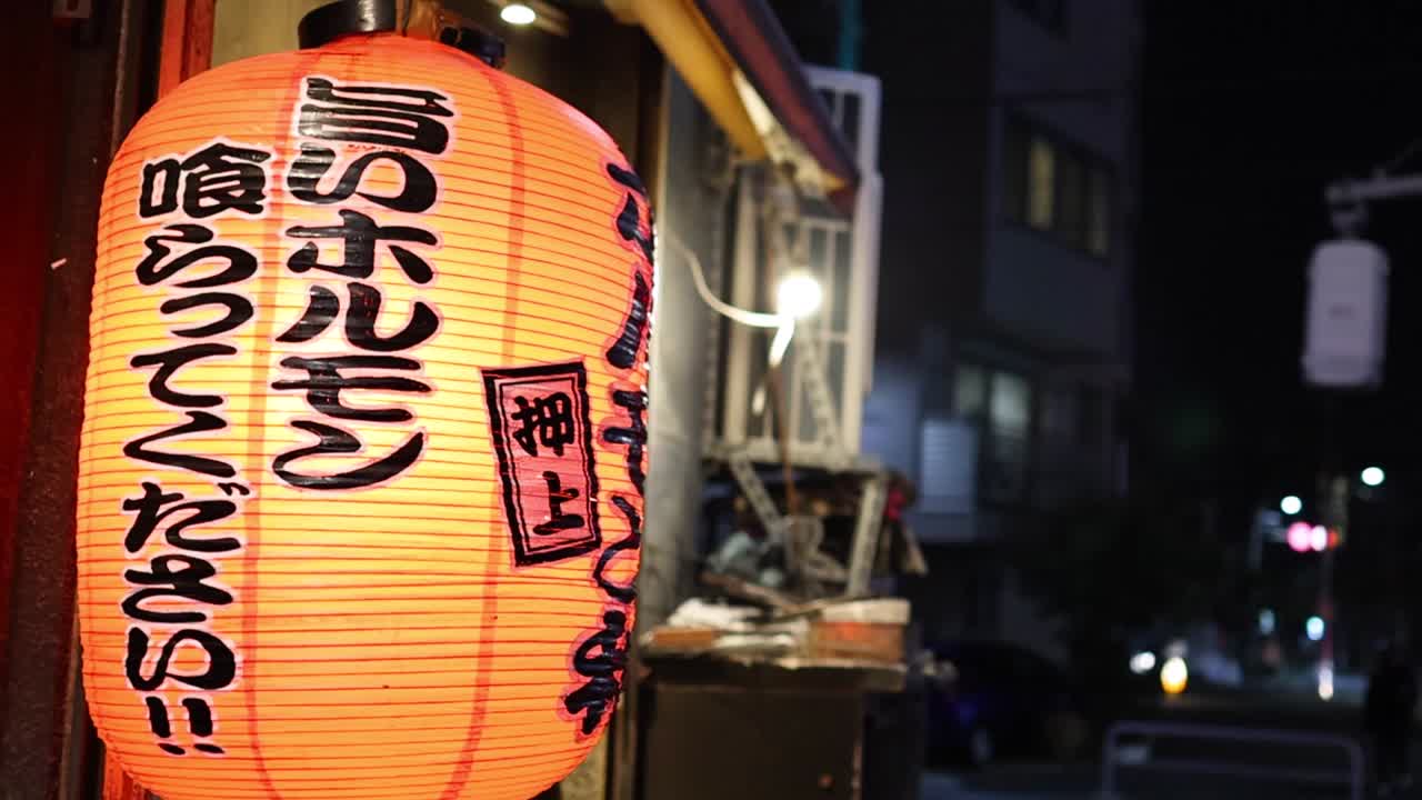 A bright lantern with Japanese characters illuminates a dimly lit urban street scene at night.