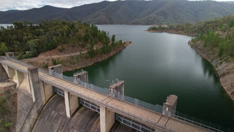 Descending-over-the-spillway-at-Lake-Eildon,-Victoria,-Australia-with-the-lake-and-mountains-behind