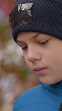 niño con gorra negra con parche y chaqueta azul paseando con la cabeza inclinada pensativamente, rodeado de borroso follaje vibrante de otoño y suave iluminación natural