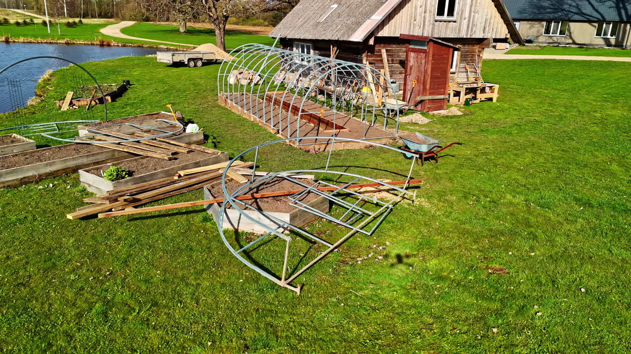 A greenhouse is being assembled in the lush green garden of a rustic wooden house.