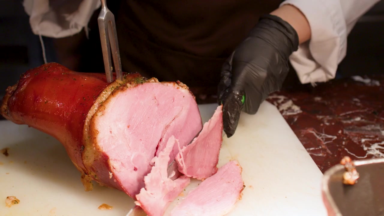 Uno chef con guanti neri usa un coltello da intaglio per affettare prosciutto arrosto al buffet.