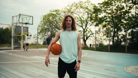 A-curly-haired-red-haired-man-in-a-light-green-T-shirt-and-with-a-basketball-in-his-hands-poses-against-the-background-of-a-team-that-plays-basketball-on-the-basketball-court