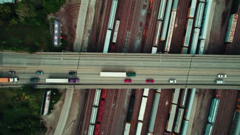 Top-down-aerial-of-green-semi-truck,-purple-tractor-and-yellow-trailer-driving-across-the-bridge-above-train-rails-network-in-Illinois-USA