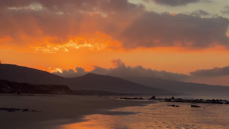 the sun shining brilliantly from behind hills during sunset with a calm beach in the foreground