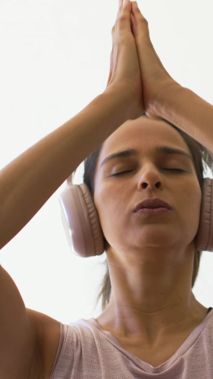 Video verticale: Donna con cuffie che guarda in alto, unisce le mani in preghiera su sfondo bianco, meditando.